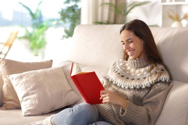 Kışın evde kanepede oturmuş kağıt kitap okuyan mutlu bir kadın.