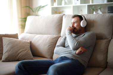 Üzgün adam evdeki bir kanepede oturmuş kulaklıktan gelen sesi dinliyor.