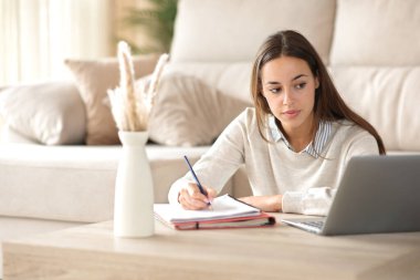 Öğrenci evde dizüstü bilgisayarda ders izlerken notlar alıyor