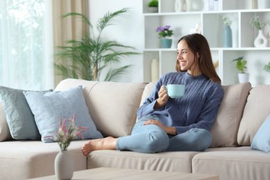 Mavi elbiseli, kahve içen mutlu bir kadın evdeki kanepede oturmuş başka tarafa bakıyor.