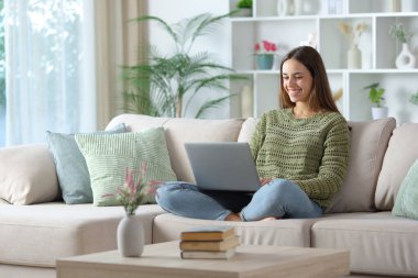 Evdeki kanepede oturan yeşil elbiseli mutlu kadın dizüstü bilgisayar kullanıyor.