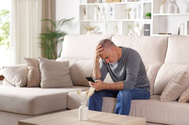 Endişeli kıdemli adam telefondaki kötü haberleri kontrol ediyor evdeki kanepede oturuyor. 