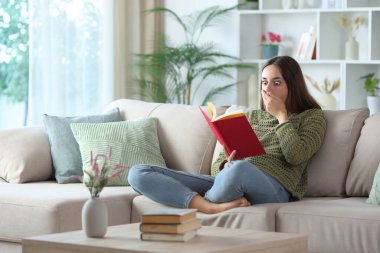 Yeşilli kadın, evdeki kanepede oturmuş kırmızı kitap okuyor.