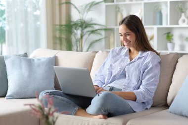 Evdeki koltukta oturan mavi elbiseli mutlu kadın dizüstü bilgisayar kullanıyor.
