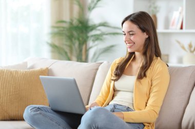 Evdeki koltukta dizüstü bilgisayar kullanan mutlu bir kadın.