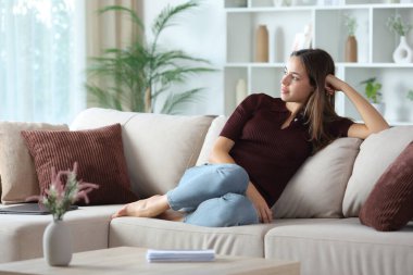 Rahat, düşünceli bir kadın evdeki bir kanepede otururken başka tarafa bakıyor.