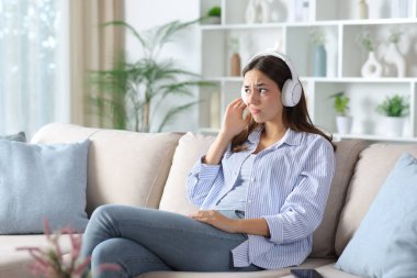 Evdeki koltukta oturan mavi elbiseli, kulaklıklı, hayal kırıklığına uğramış bir kadın.