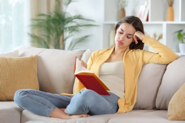 Sarı elbiseli, kırmızı kitap okuyan sıkıcı kadın evdeki kanepede oturuyor.
