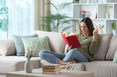 Yeşiller içindeki heyecanlı kadın evdeki kanepede oturmuş kırmızı kitap okuyor.