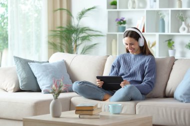 Mavi giyen, kulaklık takan, dijital tablette medyayı izleyen, evdeki koltukta oturan mutlu bir kadın.