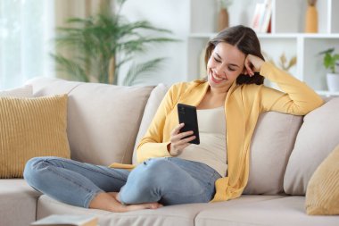 Sarı elbiseli mutlu kadın evdeki kanepede oturan akıllı telefonu kontrol ediyor.