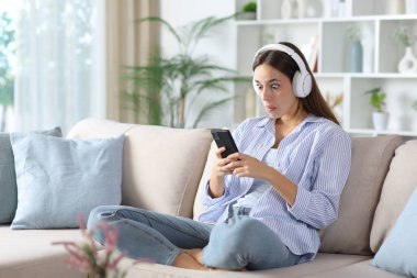 Kulaklık takmış, telefonla medyayı izleyen kafası karışmış bir kadın evdeki kanepede oturuyor.