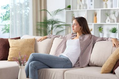 Rahatlamış bir kadın pencereden dışarı bakıyor ve evdeki kanepede yalnız başına oturduğunu düşünüyor.