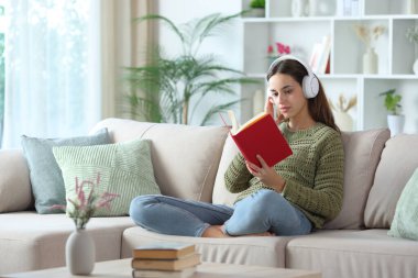 Yeşiller içinde kulaklık takıp kitap okuyan ciddi bir kadın.