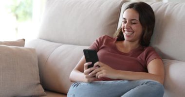 Kanepede oturup akıllı telefon kullanan ve evde gülen mutlu bir kadın.