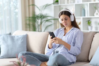 Hüsrana uğramış, mavili bir kadın telefonda medyayı izliyor. Evdeki koltukta oturuyor.