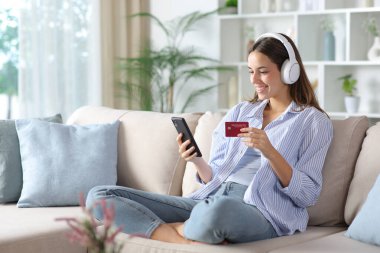 Mavili, kulaklık takan mutlu bir kadın, internetten kredi kartı ve telefonuyla evdeki bir koltukta oturuyor.