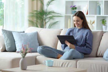 Evdeki kanepede oturan mavi elbiseli mutlu kadın tablet kullanıyor.