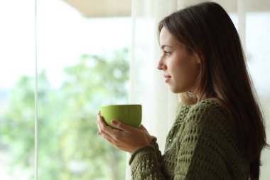 Kahve içen bir kadın evin içinde duran camdan dışarı bakıyor.