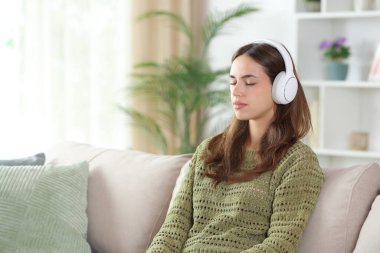 Yeşilli kadın kulaklık takıyor. Evdeki koltukta dinleniyor.
