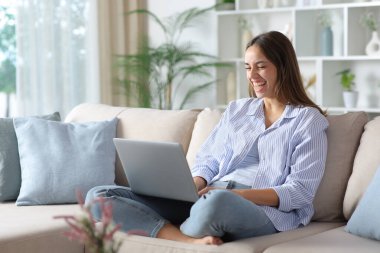 Evdeki kanepede oturup dizüstü bilgisayarın içeriğini kontrol ederken gülen mavili komik kadın.