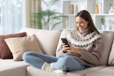 Kışın evde koltukta oturan akıllı telefonu kullanan mutlu bir kadın.
