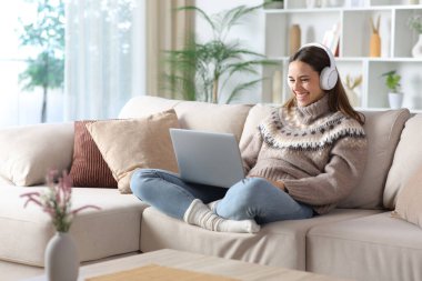 Kışın kulaklık takan mutlu bir kadın dizüstü bilgisayarı seyrediyor evdeki koltukta oturuyor.