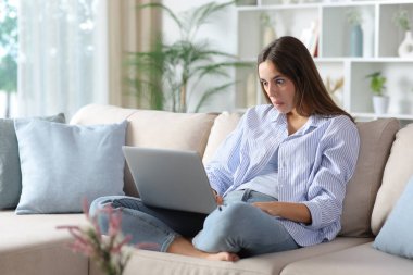 Mavili şaşkın kadın evdeki koltukta oturmuş dizüstü bilgisayarını kontrol ediyor.