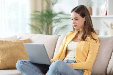 Evdeki kanepede oturup dizüstü bilgisayarını kontrol eden ciddi bir kadın.