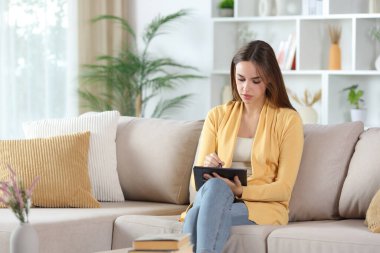 Evdeki kanepede oturan sarı el yazısıyla dijital tablet kullanan kadın.