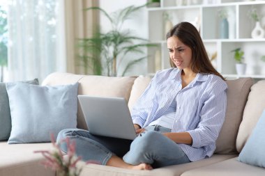 Mavili kafası karışmış bir kadın evdeki koltukta oturmuş dizüstü bilgisayarını kontrol ediyor.