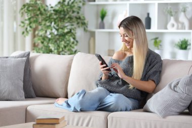 Mutlu bir kadın evdeki bir kanepede oturmuş akıllı telefonuyla internetten içeriklerini karıştırıyor.