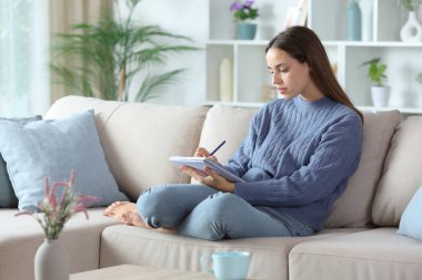 Serious woman in blue writing notes on notebook sitting on a couch at home