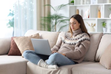 Ciddi bir kadın kışın dizüstü bilgisayarla evde bir kanepede oturuyor.