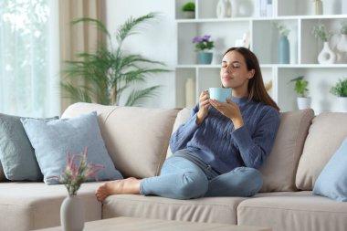 Woman in blue smelling coffee and relaxing sitting on a couch at home