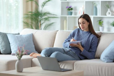 Woman in blue taking notes of laptop online content sitting on a sofa at home