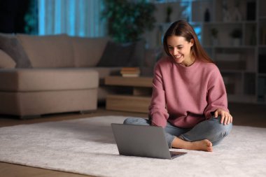 Happy woman in the night using laptop sitting on a carpet at home