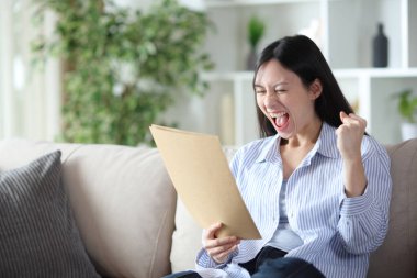 Heyecanlı Asyalı kadın evdeki koltukta oturmuş belgeleri kontrol ediyor.