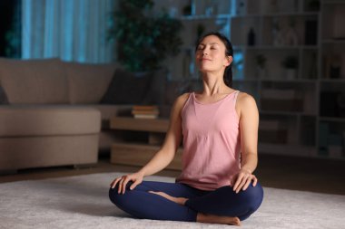 Gece yoga yapan Asyalı bir kadın evde halının üzerinde oturmuş nefes alma egzersizi yapıyor.