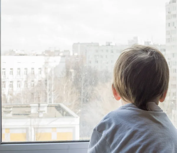 A little boy looks out the window. The street is bright and sunny. Cold weather — Stock Photo, Image