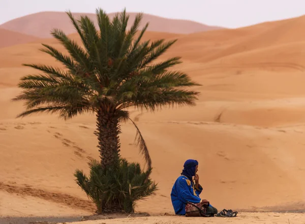 青い衣を着た男がサハラ砂漠の砂のヤシの木の下に座り — ストック写真