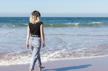 Genç bir kadın bir yaz günü kot pantolon ve siyah bluzla Sopelan sahilinde yürüyor.