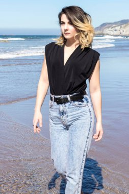 Genç bir kadın bir yaz günü kot pantolon ve siyah bluzla Sopelan sahilinde yürüyor.