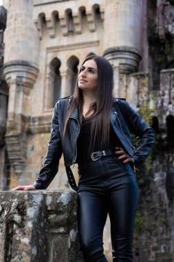 Eski bir şatonun önünde deri ceket giymiş güzel esmer bir kadının portresi.