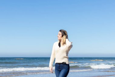 Genç bir kadın güneşli bir günde kumsalda yürüyor, kot pantolon ve yün bir kazak giyiyor.