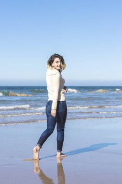 Genç bir kadın güneşli bir günde kumsalda yürüyor, kot pantolon ve yün bir kazak giyiyor.