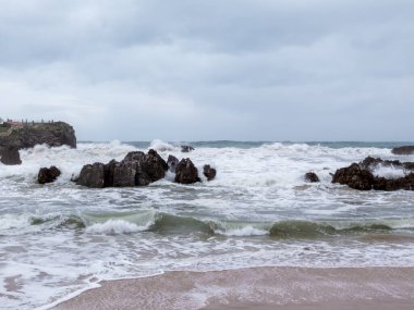 Asterias sahillerinde bulutlu bir günde kara kayaların üzerinden dalgalar kırılıyor.