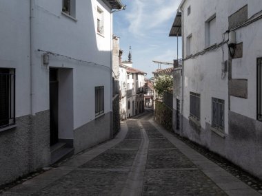 Dar kaldırımlı cadde yokuş aşağı iniyor, berrak gökyüzünün altında geleneksel beyaz sıva binalar var.