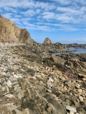Kayalık bir plaj, dramatik uçurumlar ve beyaz bulutlu mavi gökyüzü içeren engebeli kıyı şeridi