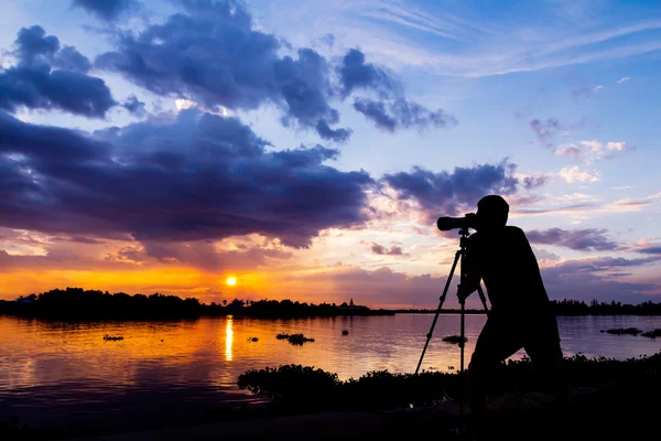 Silhueta de fotógrafo tirar foto ao pôr do sol ao lado do riv Imagens De Bancos De Imagens